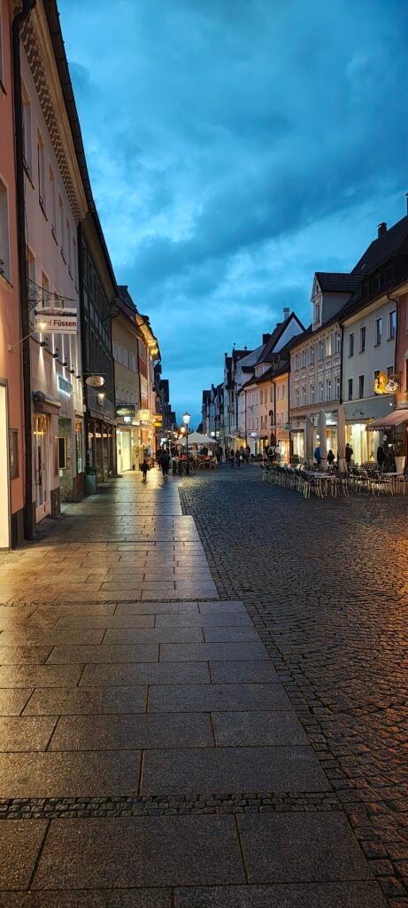 Rua de paralelepípedo no centro histórico de Füssen ao entardecer, com cafés, mesas externas e casas coloridas na Baviera, Alemanha.